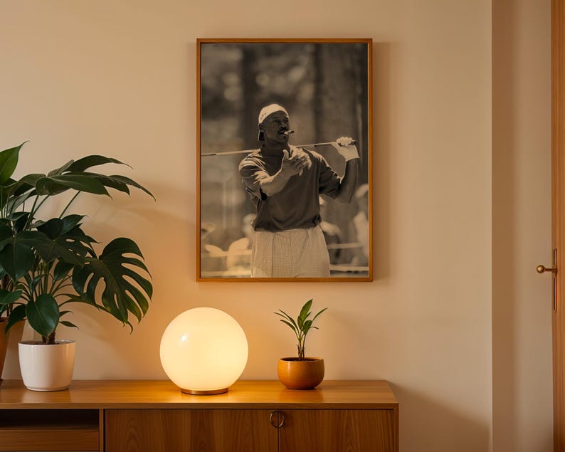 May include: A framed black and white photograph of a golfer in action, wearing a cap and holding a golf club. The photograph is displayed on a light-colored wall above a wooden cabinet. A spherical lamp and potted plants are in the foreground.