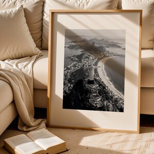 May include: A framed monochrome photograph of a coastal city, featuring a mountain with a statue, a beach, and buildings. The photograph is displayed in a light-coloured wooden frame, leaning against a beige sofa. An open book rests on the floor.