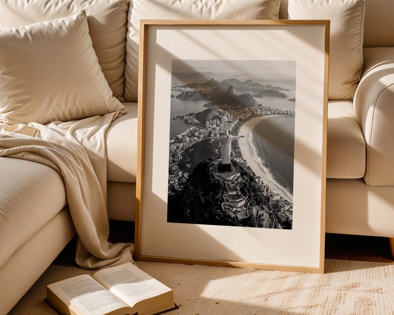 May include: A framed monochrome photograph of a coastal city, featuring a mountain with a statue, a beach, and buildings. The photograph is displayed in a light-coloured wooden frame, leaning against a beige sofa. An open book rests on the floor.