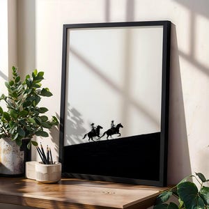 May include: Framed black and white art print featuring two silhouetted cowboys riding horses across a black hill against a white background. The artwork is displayed in a black frame and is set on a wooden surface.