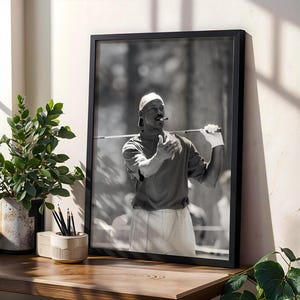 以下が含まれることがあります： ゴルフのクラブと葉巻を持っている人の白黒の額入り写真。その人は白い帽子、半袖シャツ、白い手袋を着用しています。写真は植木鉢の隣の木製の表面に展示されています。