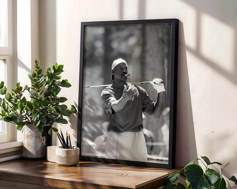 May include: Black and white framed photograph of a person holding a golf club and a cigar. The person is wearing a white cap, a short-sleeved shirt, and white gloves. The photograph is displayed on a wooden surface next to a potted plant.