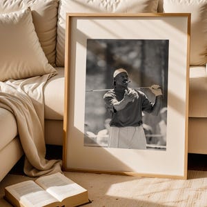May include: Black and white framed photograph of a person playing golf. The person is wearing a cap, polo shirt, and shorts, holding a golf club. The photograph is in a wooden frame, leaning against a beige couch.