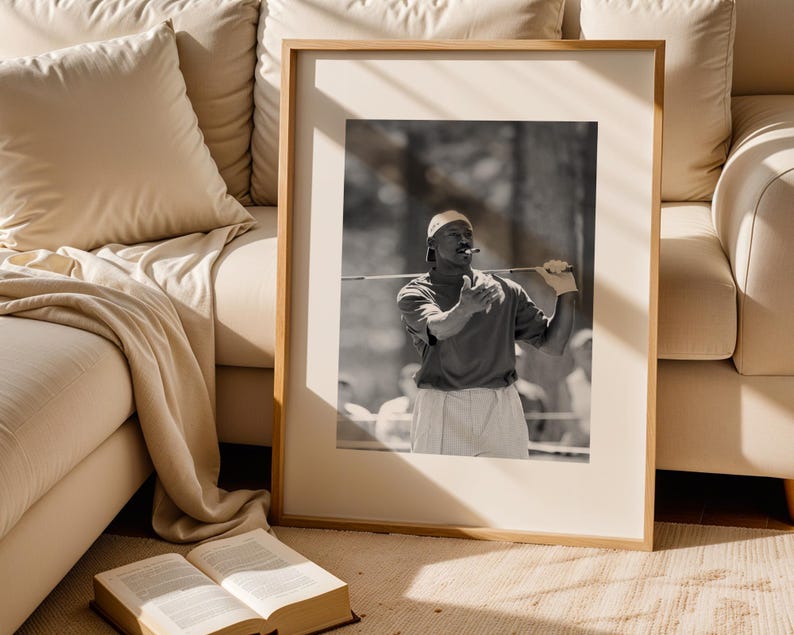 May include: Black and white framed photograph of a person playing golf. The person is wearing a cap, polo shirt, and shorts, holding a golf club. The photograph is in a wooden frame, leaning against a beige couch.