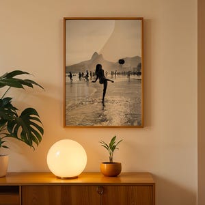 May include: A framed sepia-toned photograph depicts a person kicking a ball on a beach, with a mountain in the background. A round, glowing lamp sits on a wooden cabinet, next to a potted plant. The image is displayed on a beige wall.