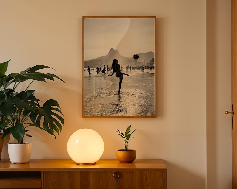 May include: A framed sepia-toned photograph depicts a person kicking a ball on a beach, with a mountain in the background. A round, glowing lamp sits on a wooden cabinet, next to a potted plant. The image is displayed on a beige wall.