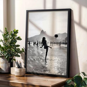 May include: Black and white framed print of a beach scene. A person is kicking a soccer ball on the beach, with other people in the background. Mountains are visible in the distance. The print is displayed on a wooden surface with a plant and pencils.
