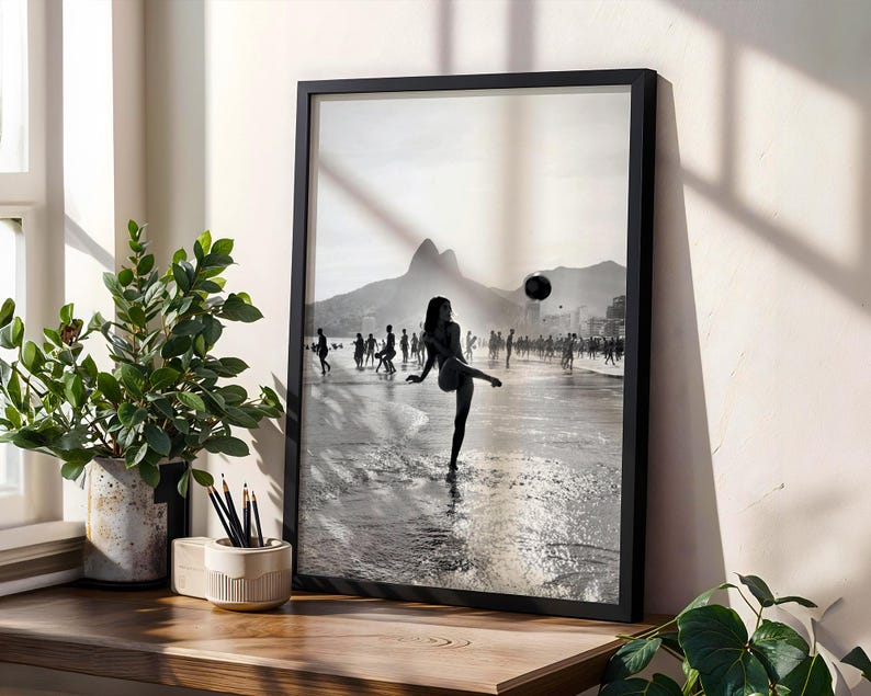May include: Black and white framed print of a beach scene. A person is kicking a soccer ball on the beach, with other people in the background. Mountains are visible in the distance. The print is displayed on a wooden surface with a plant and pencils.