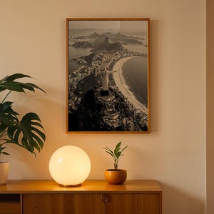 May include: A framed monochrome photograph of a coastal city scene hangs on a beige wall. A glowing spherical lamp sits on a wooden cabinet, with a potted plant to the left and a small plant in an orange pot to the right.