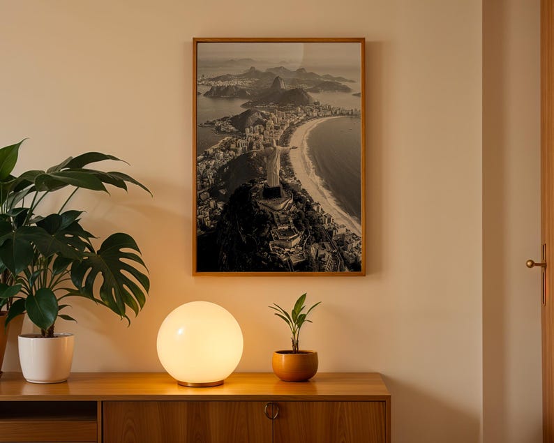 May include: A framed monochrome photograph of a coastal city scene hangs on a beige wall. A glowing spherical lamp sits on a wooden cabinet, with a potted plant to the left and a small plant in an orange pot to the right.