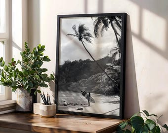 May include: Black and white framed print of a tropical beach scene. A surfer stands on the sand holding a surfboard. Palm trees and lush vegetation are visible in the background. The print is displayed on a wooden surface with a potted plant and pencils.