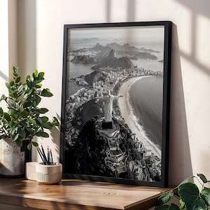 May include: Monochrome framed print of Rio de Janeiro, Brazil, showing the Christ the Redeemer statue on Corcovado Mountain, with the city and coastline. The print is on a wooden surface, with a plant and pencil holder.