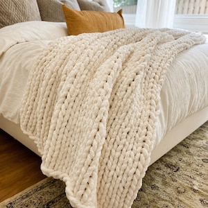 May include: A chunky knit throw blanket in an off-white cream color. The blanket features a thick, hand-knitted texture with large, interconnected loops of soft yarn, draped over the edge of a bed with a smooth, solid-colored white bedspread.