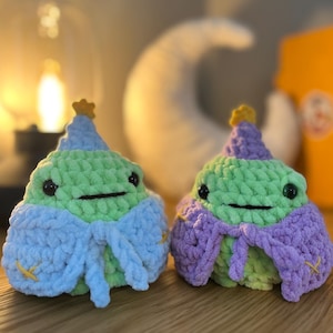 May include: Two handmade crochet plushies shaped like rounded creatures with light green bodies, black bead eyes, and small stitched mouths. Each wears a pointed hat topped with a yellow star and a matching cape tied at the front, one in light blue and one in purple.