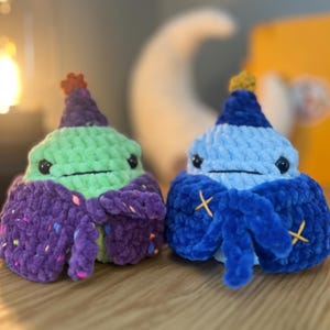 Puede incluir: Dos criaturas de ganchillo de felpa con cuerpos redondos y sombreros puntiagudos. La criatura de la izquierda es verde claro con una capa morada con motas de colores. La de la derecha es azul claro con una capa azul oscuro y detalles en cruz amarillos.