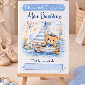 Peut inclure: Un livret d'activités de baptême sur le thème marin. La couverture présente un ours de dessin animé portant un chapeau de marin naviguant sur un bateau, avec le texte "Petit carnet d'activités" et "Mon Baptême". Le livret est présenté sur un petit chevalet en bois.