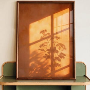 Puede incluir: Una obra de arte enmarcada con un fondo cálido y una silueta de planta. El marco de madera rodea la obra, y la sombra de una ventana se proyecta sobre ella. La obra se exhibe sobre una estantería verde.