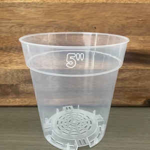 May include: A clear, semi-transparent plastic plant pot featuring a wide rim and a perforated base for drainage. The upper portion of the pot is printed with the text 5 inch, indicating its size. The design includes a raised, circular grid pattern at the bottom to facilitate airflow and water movement for plant roots.