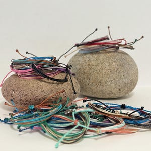 May include: A collection of colorful string bracelets in various shades of blue, pink, green, and orange. The bracelets are arranged on and around two smooth, gray stones. The bracelets have adjustable knots.