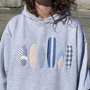 May include: A pale grey hoodie with a design of several surfboards in different patterns and colours. The surfboards include stripes, gingham, and solid colours, with a light pink surfboard in the centre. The hoodie has a drawstring hood.
