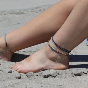 May include: Anklets on a person's ankles. One ankle has a single green and red beaded anklet. The other ankle has three anklets in shades of brown and blue. The person is sitting on a sandy beach.