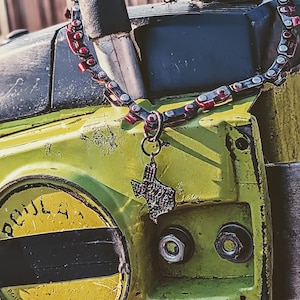 May include: A necklace made from a chainsaw chain with red and silver links, and a Texas-shaped pendant. The necklace hangs over a vintage, yellow and black chainsaw, with the word "POULAN" visible on the side.