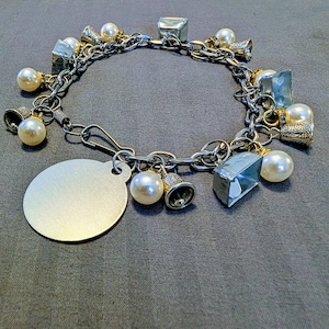 May include: A silver charm bracelet with various charms, including faux pearls, small bells, and geometric shapes. The bracelet has a chain link design and a round, blank tag. The charms are in silver and gold tones.
