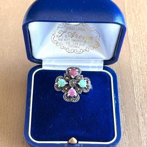 May include: A silver-toned ring featuring a four-leaf clover design with two pink pear-shaped stones, two round turquoise-colored stones, and small dark grey accent stones, presented in a blue velvet-lined jewelry box with gold text.