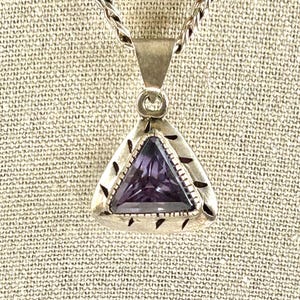 May include: A triangular pendant featuring a faceted purple gemstone set in a silver frame with decorative etched markings along the edges. The pendant hangs from a silver rope chain, showcasing a polished metallic finish and a clear, deep violet stone.