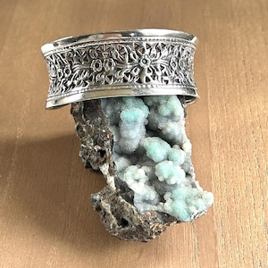 May include: A wide silver cuff bracelet featuring an intricate embossed floral and leaf pattern along its band, resting atop a rough, dark brown mineral specimen covered in clusters of small, light mint-green crystalline formations.
