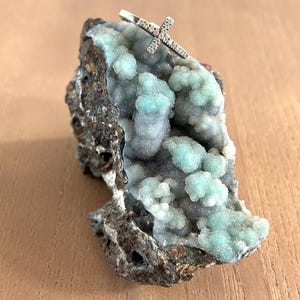 May include: A silver ring with a cross design, encrusted with small, sparkling stones. The ring rests on a piece of rough, textured rock with a mix of gray, brown, and light blue-green colors. The background is a light brown wooden surface.