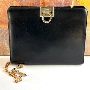 May include: A black rectangular leather handbag featuring a gold-tone metal Gancini clasp closure at the top center. The clasp is engraved with the Ferragamo brand name. A gold-tone chain strap is attached to the side of the structured bag.