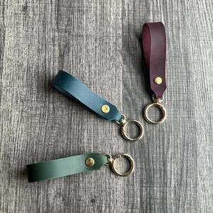 May include: Three leather keychains in teal, burgundy, and green. Each keychain has a gold-colored metal ring and a brass rivet. The keychains are displayed on a wooden surface.