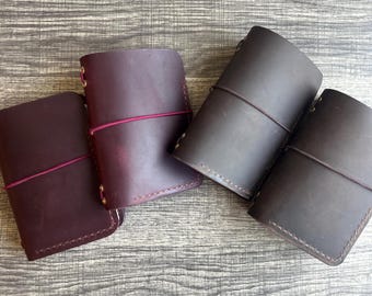 May include: Four leather journals in shades of burgundy and brown. Each journal is closed with an elastic band and features gold-colored rivets. The journals are arranged on a wooden surface.