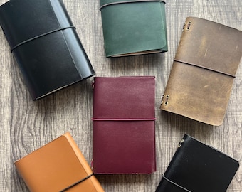Può includere: Una collezione di diari in pelle in vari colori, tra cui nero, verde, marrone, bordeaux e marrone chiaro. Ogni diario è chiuso con un elastico. I diari sono disposti su una superficie di legno.