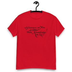 May include: A red t-shirt with the quote "Ignorance on fire is better than knowledge on ice" in black script. The shirt is hanging from a black hanger.