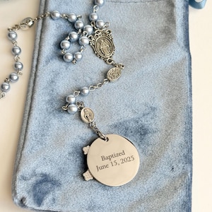 May include: A rosary featuring light blue beads and silver-toned metal links, including a central medal and a circular pendant engraved with the text Baptized June 15, 2025. The rosary is displayed on a light blue velvet drawstring pouch.