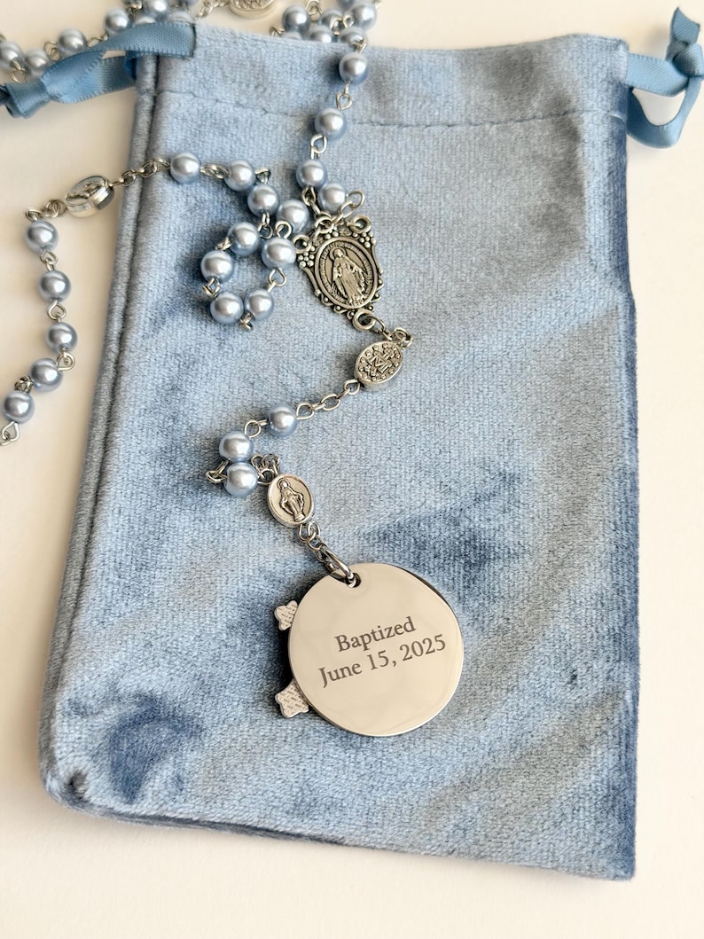 May include: A rosary featuring light blue beads and silver-toned metal links, including a central medal and a circular pendant engraved with the text Baptized June 15, 2025. The rosary is displayed on a light blue velvet drawstring pouch.