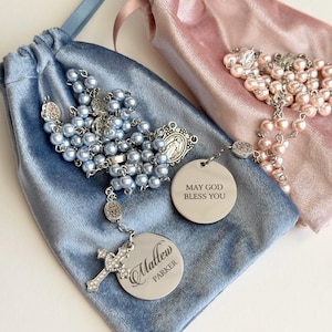 May include: Two rosaries with light blue and light pink pearl beads, silver-tone metal links, and crucifixes. One rosary features a round silver charm engraved with 'Matthew Parker' and a cross, while the other has a charm reading 'MAY GOD BLESS YOU'. Both are displayed on matching velvet drawstring pouches.