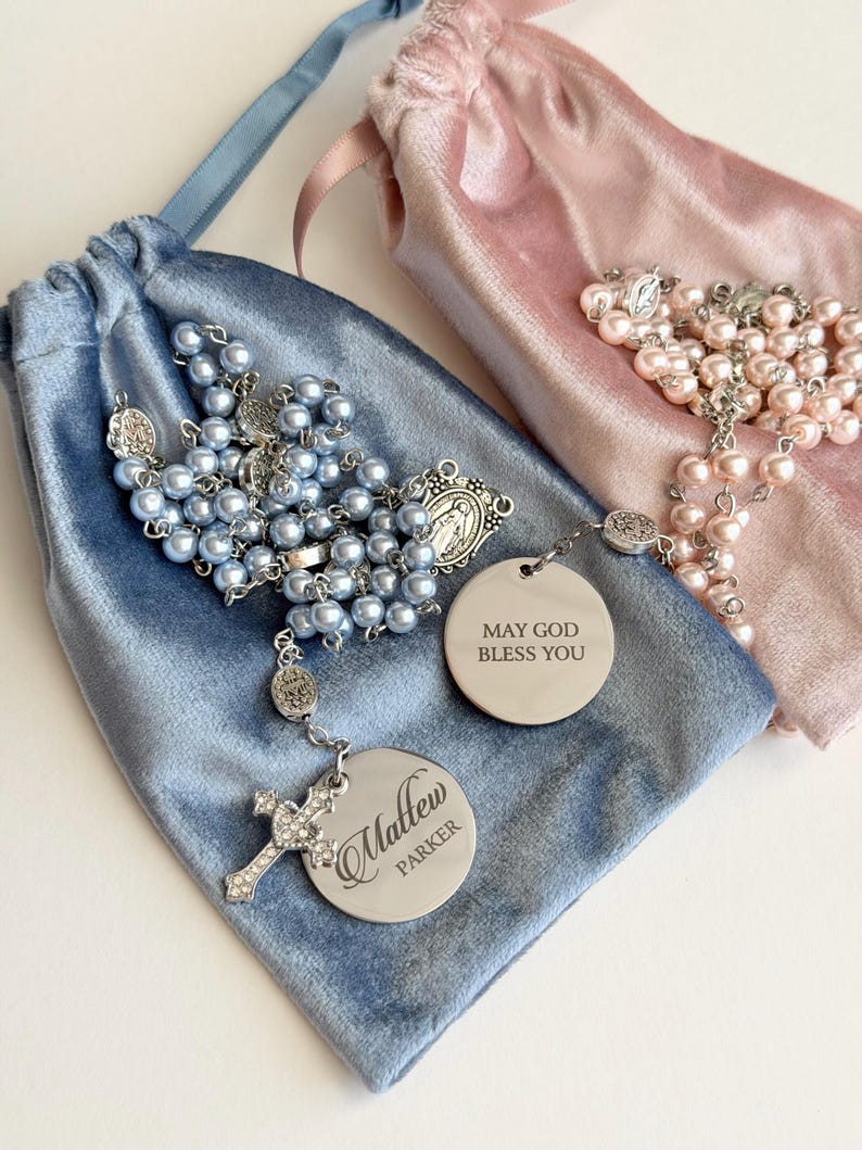 May include: Two rosaries with light blue and light pink pearl beads, silver-tone metal links, and crucifixes. One rosary features a round silver charm engraved with 'Matthew Parker' and a cross, while the other has a charm reading 'MAY GOD BLESS YOU'. Both are displayed on matching velvet drawstring pouches.