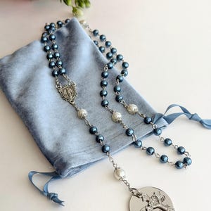 May include: A rosary featuring dark blue and white pearl-like beads on a silver-toned chain, with a central silver medal depicting a figure, and a circular silver pendant with a crystal-encrusted cross and the partial text "PAR" engraved.