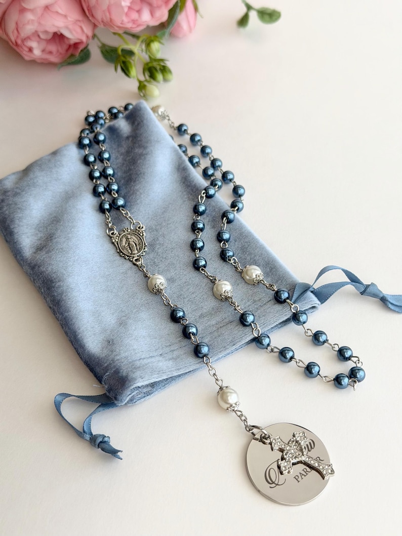 May include: A rosary featuring dark blue and white pearl-like beads on a silver-toned chain, with a central silver medal depicting a figure, and a circular silver pendant with a crystal-encrusted cross and the partial text "PAR" engraved.