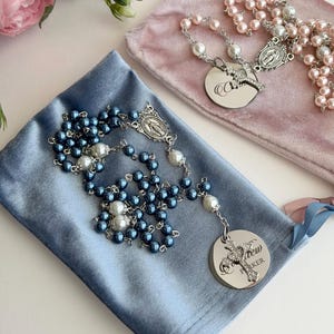 May include: Two rosaries featuring metallic blue and soft pink pearl beads, silver-toned metal chains, and oval religious medallions. Each rosary includes a silver cross charm and a circular metal pendant engraved with the name Parker.