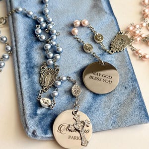 May include: Two rosaries with light blue and light pink pearl-like beads on silver-tone chains. One rosary features a round silver-tone pendant engraved with the text MAY GOD BLESS YOU, while the other has a round pendant with engraved text.