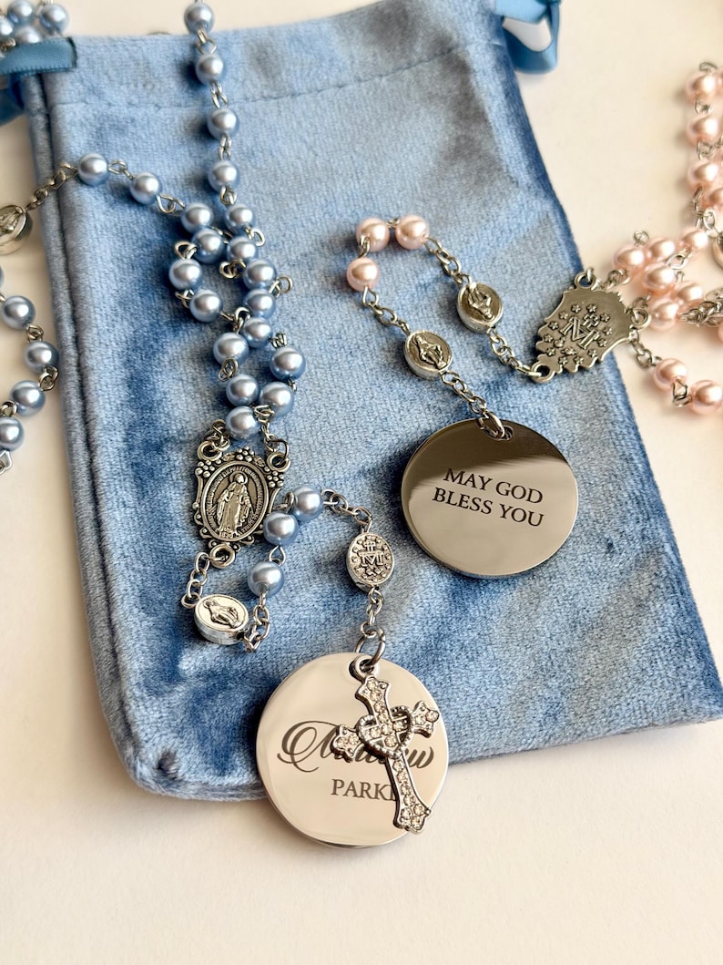 May include: Two rosaries with light blue and light pink pearl-like beads on silver-tone chains. One rosary features a round silver-tone pendant engraved with the text MAY GOD BLESS YOU, while the other has a round pendant with engraved text.