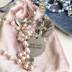 May include: A rosary featuring light pink pearl-like beads, a silver-toned metal cross, and a circular silver charm engraved with the text MAY GOD BLESS YOU. A second silver charm is engraved with the name Parker. A blue velvet pouch is visible.
