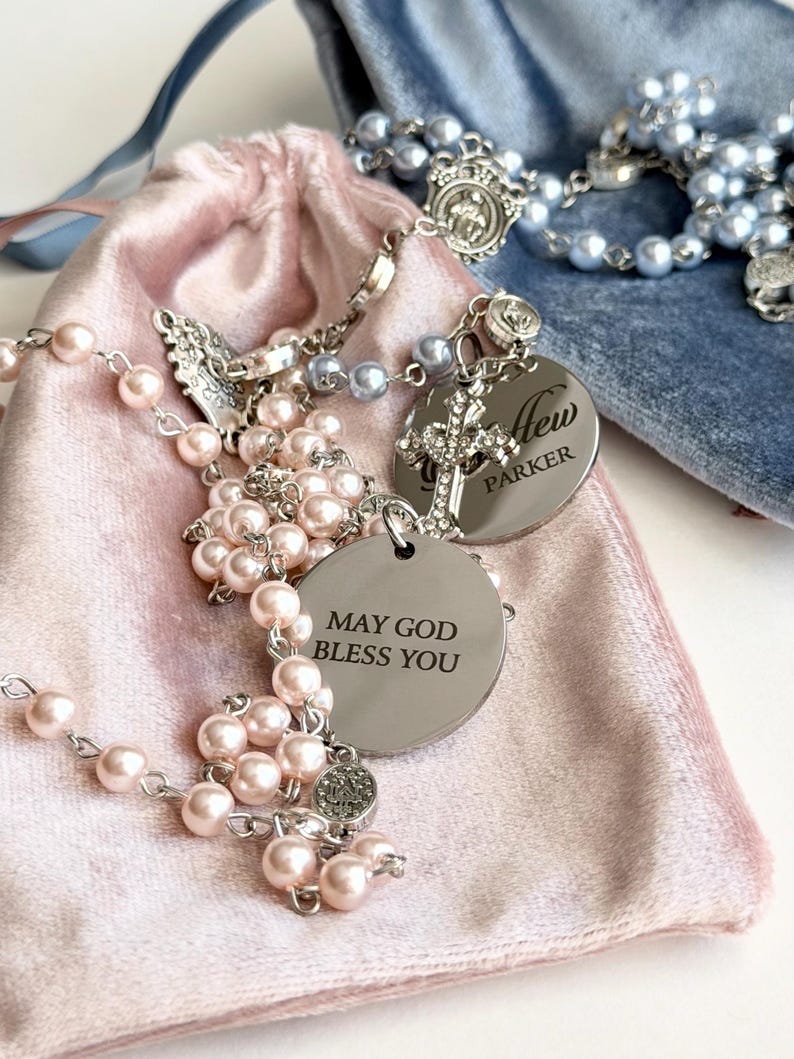 May include: A rosary featuring light pink pearl-like beads, a silver-toned metal cross, and a circular silver charm engraved with the text MAY GOD BLESS YOU. A second silver charm is engraved with the name Parker. A blue velvet pouch is visible.