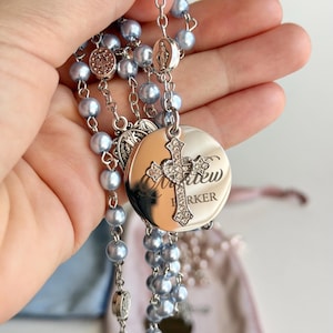 May include: A rosary featuring light blue pearl-like beads connected by silver-toned metal links. It includes a circular silver-toned pendant engraved with the name Matthew Barker, topped with a small, ornate cross encrusted with clear crystals.