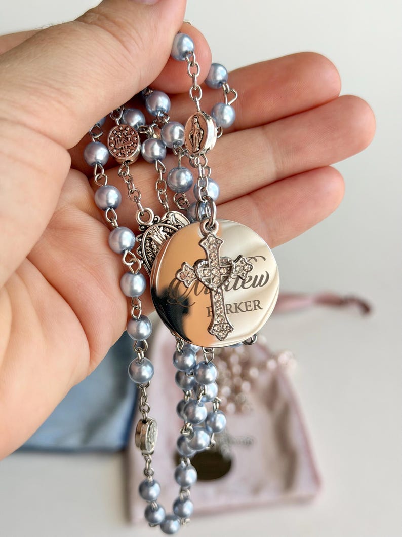 May include: A rosary featuring light blue pearl-like beads connected by silver-toned metal links. It includes a circular silver-toned pendant engraved with the name Matthew Barker, topped with a small, ornate cross encrusted with clear crystals.
