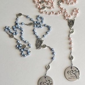 May include: Two rosaries featuring light blue and light pink pearl-like beads on silver-toned metal chains. Each rosary includes a central religious medal and a circular silver pendant engraved with text and a small rhinestone-encrusted cross charm.